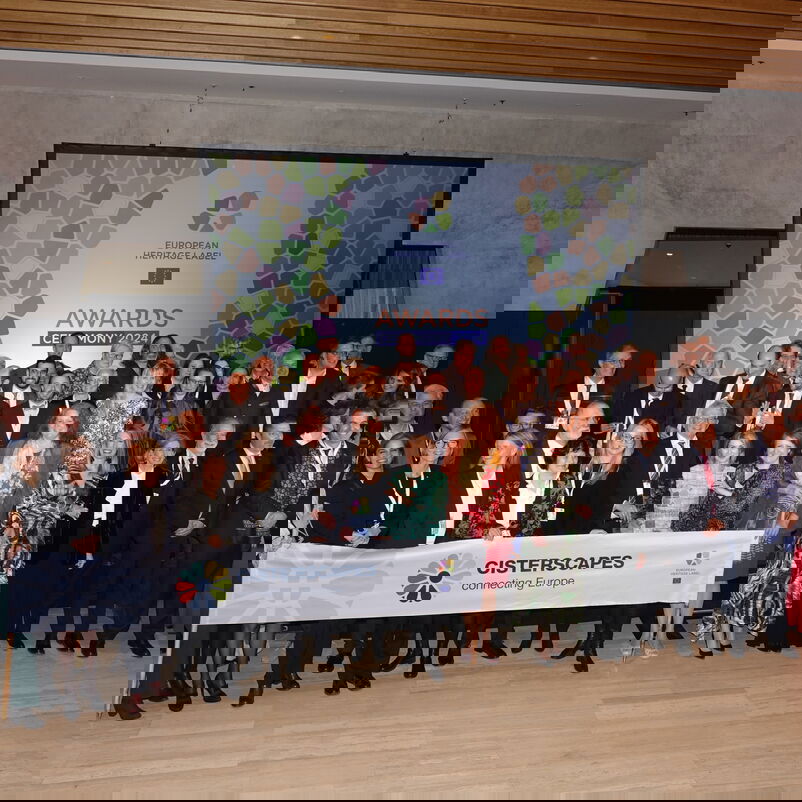 Eine Gruppe von weiblich und männlich zu lesenden Personen in festlicher Kleidung stehen in einem Halbrund und halten ein Transparent hoch, das auf ausgezeichnete Netzwerk verweist.