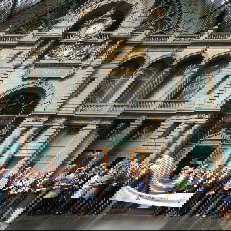 Weiblich und männlich zu lesende Personen stehen in einem Halbkreis vor einem Gebäude und halten ein Transparent.