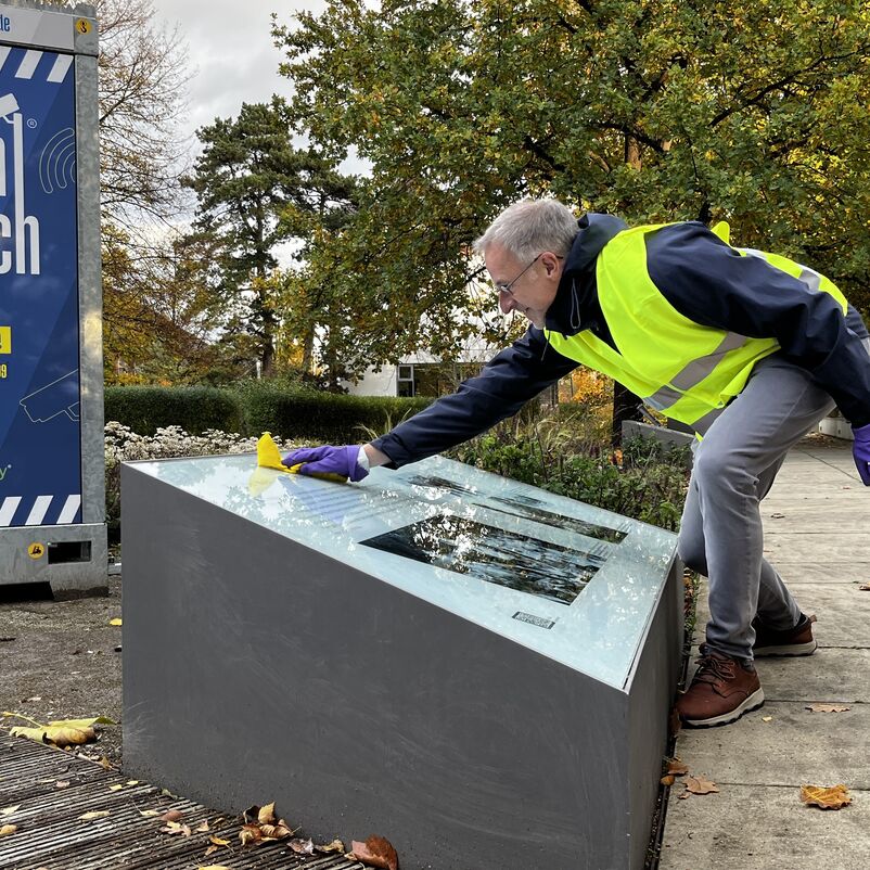 Eine männlich gelesene Person in Jacke und Warnweste putzt mit einem Wischlappen eine Schautafel in einem Garten