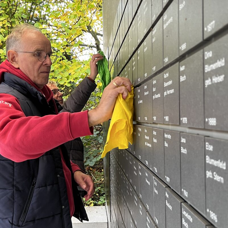 eine männlich gelesene Person putzt im Außenbereich mit einem Wischlappen Namenstafeln an einer Wand.