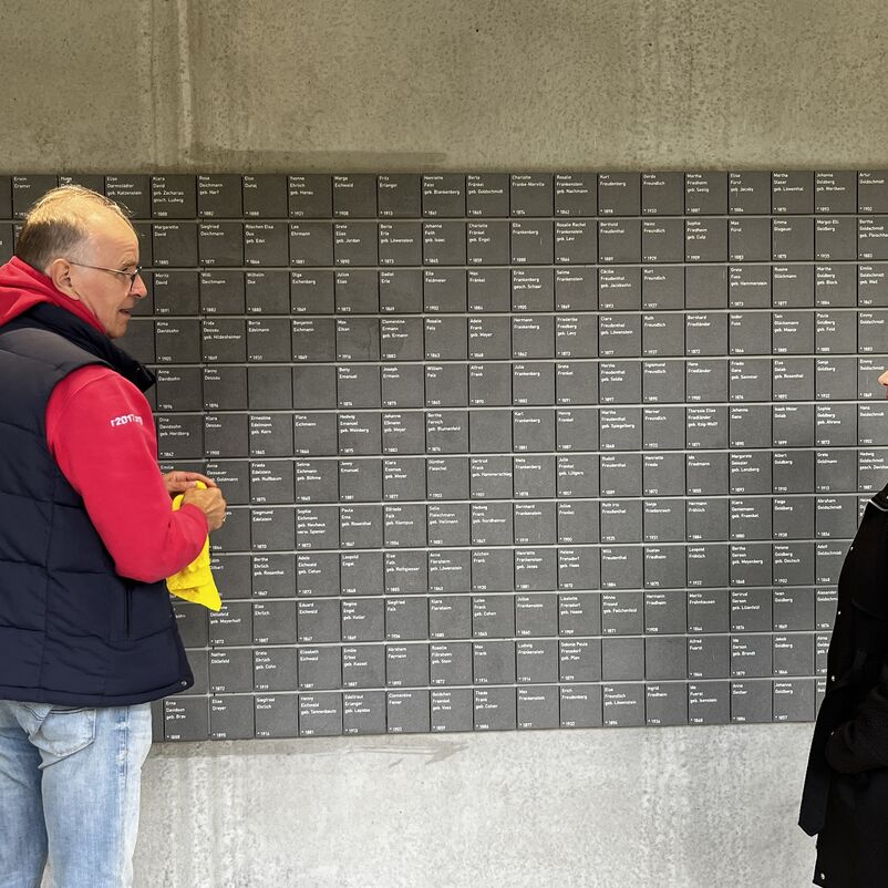 eine männlich und eine weiblich gelesene Person im Gespräch vor einer Wand mit Namenstafeln