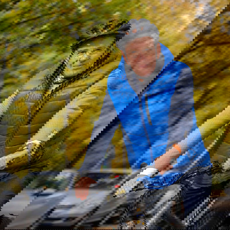 Das Bild zeigt eine männlich lesbare Person auf einem Fahrrad sitzend. Die Person trägt eine blaue Stepp-Weste, eine Fahrradhelm und eine Brille. 