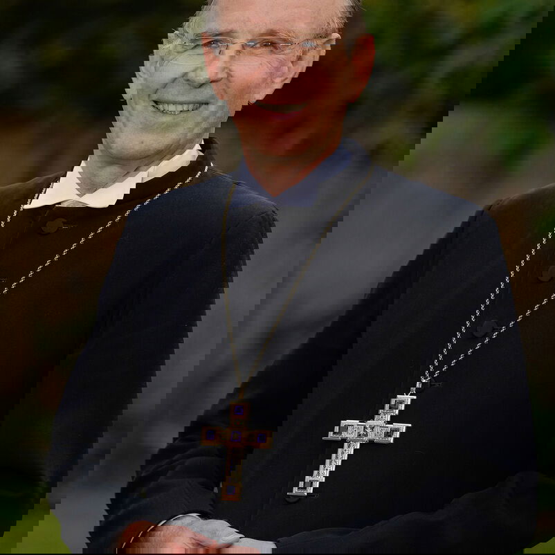 Das Bild zeigt eine männlich lesbare Person mit schwarzem Talar und einer Kette mit einem größeren goldenen Kreuz. Die Person steht auf einer Rasenfläche, lächelt, hat kurze helle Haare und eine Brille.