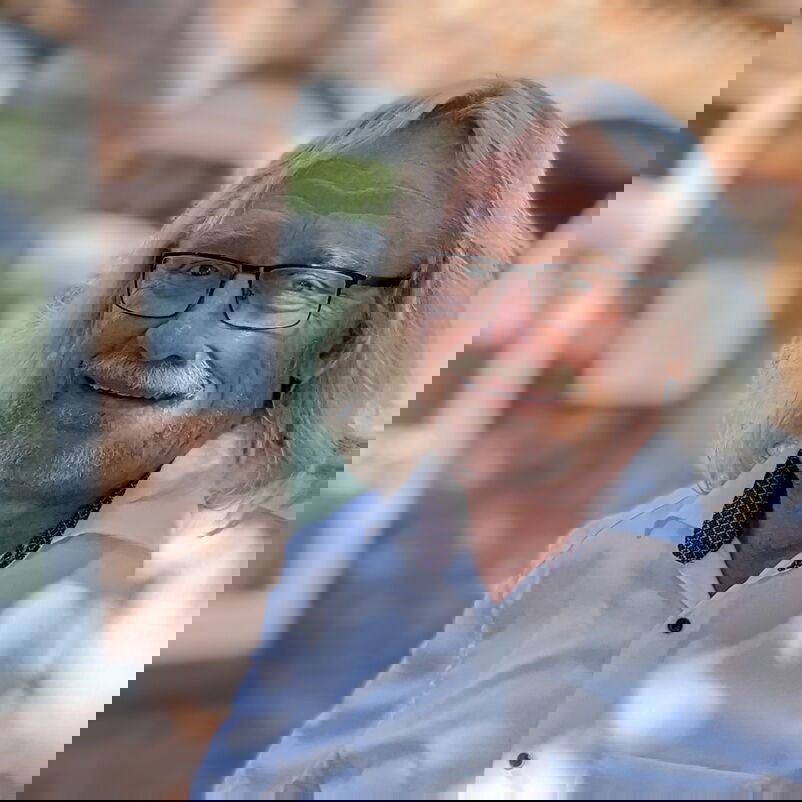 Ein weißhaariger Mann mit Brille und Hemd steht vor einem Backstein-Haus mit weißen Fensterrahmen.