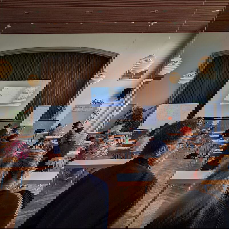 Ein Mann und eine Frau sind von hinten zu sehen, wie sie in einen großen Raum mit Holzdecke treten, an der rechten Seite eine große Fensterfront. Im Raum verteilt sitzen Menschen an Tischen.