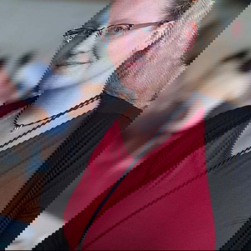 Eine blonde Frau mit Brille in rotem Shirt und schwarzer Jacke lächelt in die Kamera.