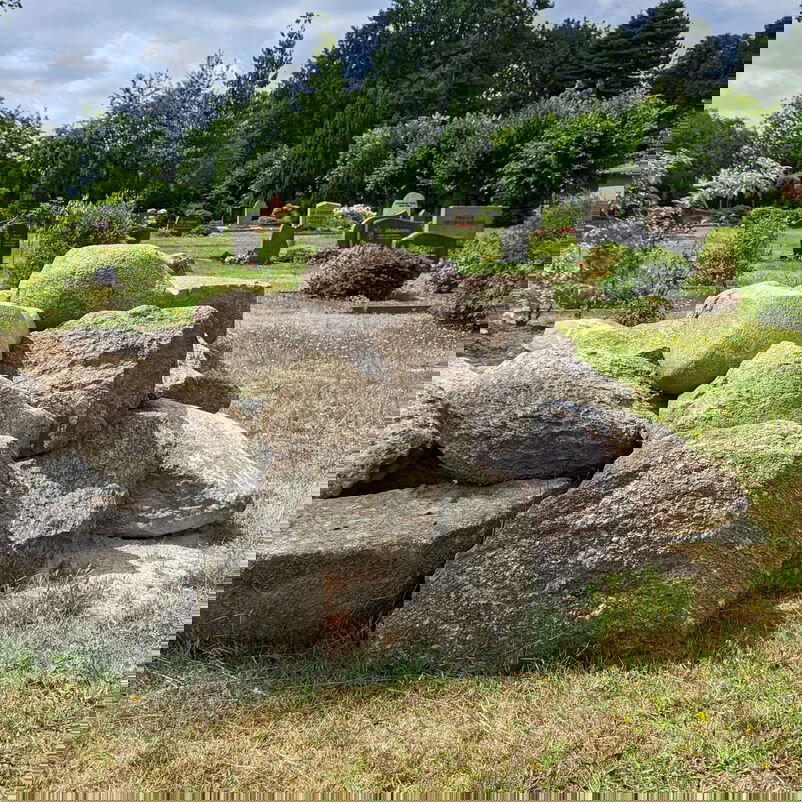 Steine sind zu einem großen Haufen geschichtet, im Hintergrund sind Gräber eines Friedhofs zu erkennen.