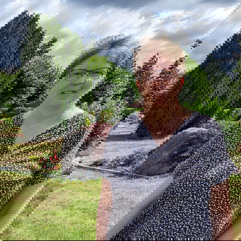 Eine Frau mit Brille und blau-weißer Bluse steht auf einem Friedhof.