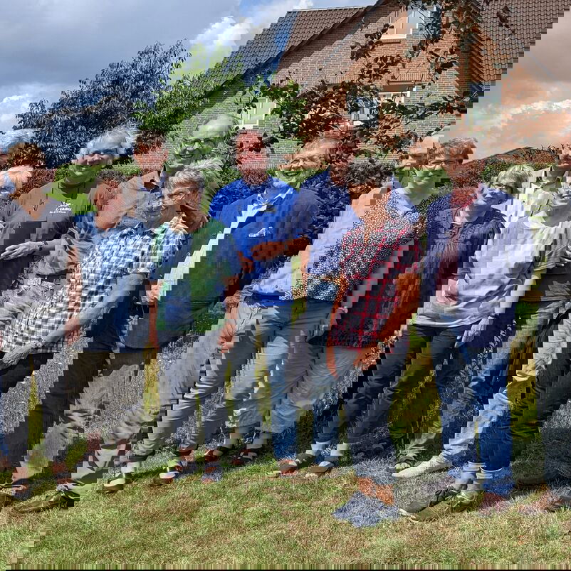 Mehrere Menschen stehen zusammen auf einer grünen Fläche, im Hintergrund Backstein-Einfamilienhäuser.