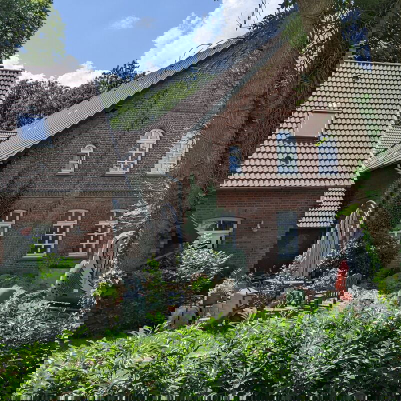 Ein großes Haus, fast ein kleiner Hof ist über eine grüne Hecke zu sehen, rechts halb verdeckt von einem Baum.