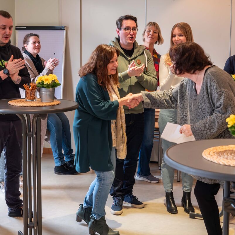 Mehrere Personen stehen an Stehtischen, klatschen und blicken fröhlich. Zwei Personen reichen sich die Hand.