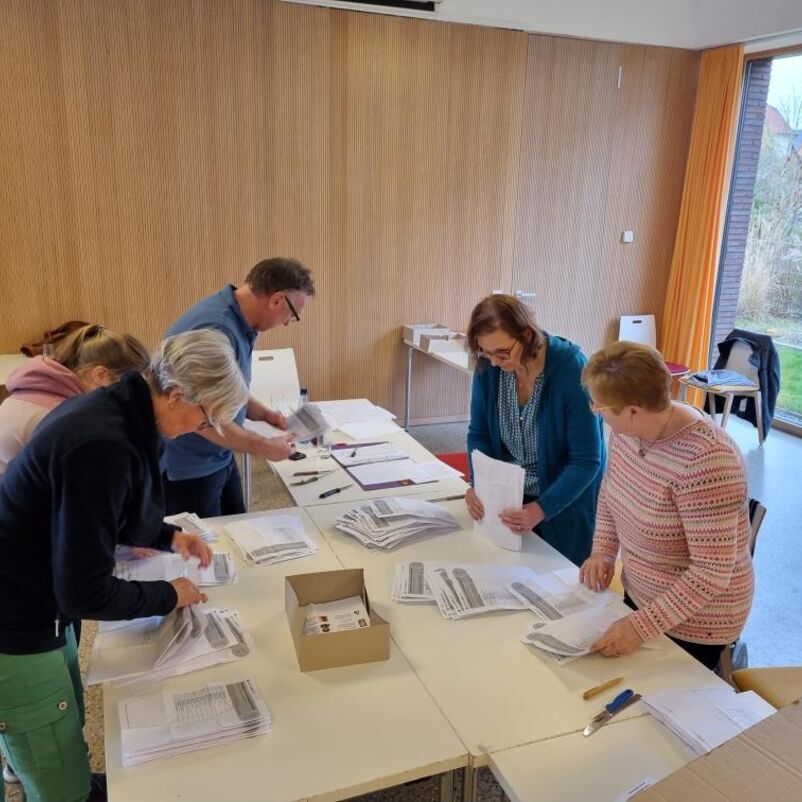 Fünf weiblich gelesene Personen beugen sich über einen Tisch, auf dem Zettel in verschiedenen Stapeln liegen.