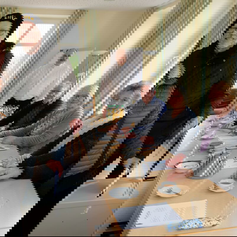 Zwei Personen stehen links von einer Tischreihe, eine von ihnen steckt einen Zettel in eine graue Wahlurne. Rechts vom Tisch sitzen mehrere Personen und sehen zu.