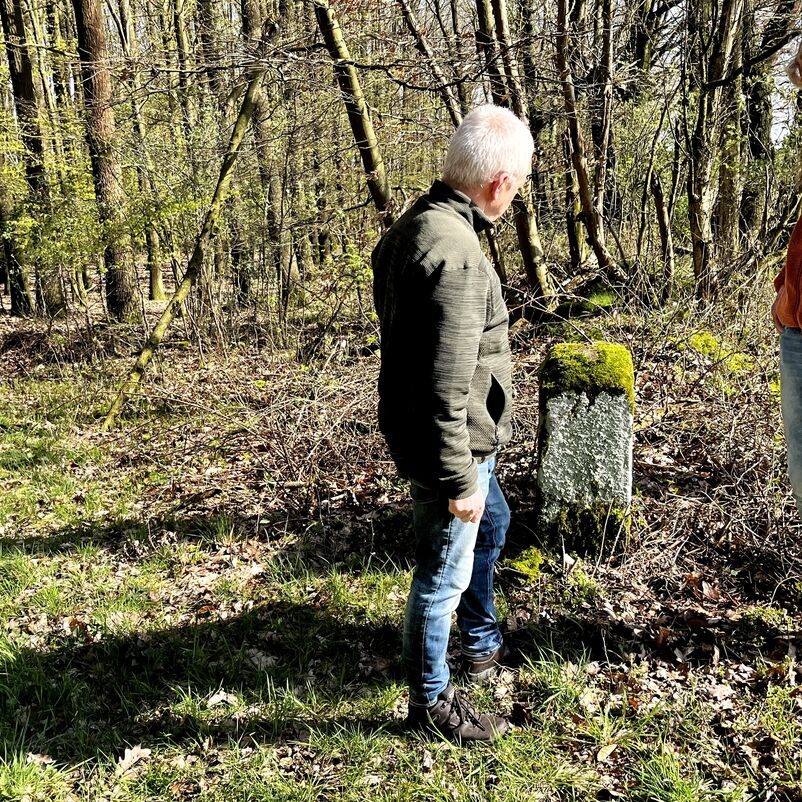 Eine männlich gelesene und eine weiblich gelesene Person stehen am Waldrand vor einem moosüberwuchterten Grenzstein. 