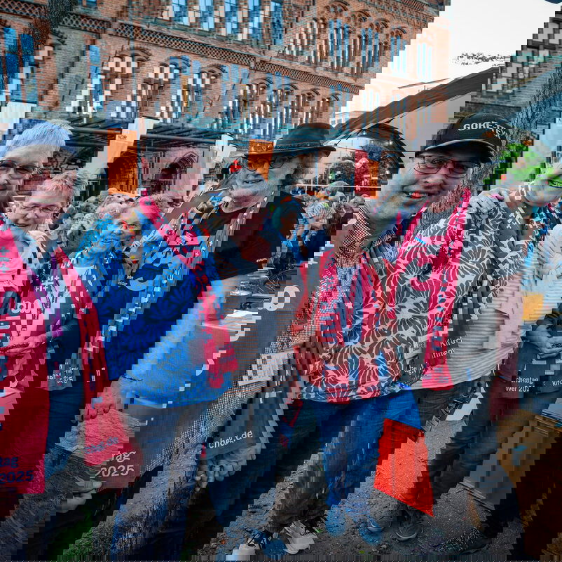 Weiblich und männlich zu lesende Personen stehen Arm in Arm auf einem Platz.