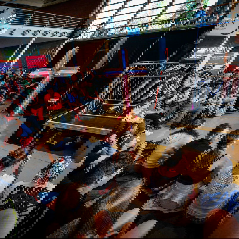 Eine Gruppe von Personen sitzt in Stuhlreihen und schaut auf eine Person, die hinter einem DJ-Pult steht.