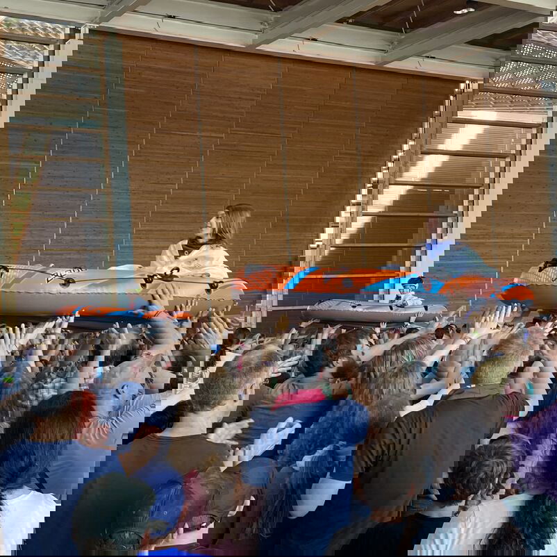 Eine Gruppe von Personen hebt in einer Halle ein Schlauchboot hoch über die Köpfe, in dem eine Person sitzt.