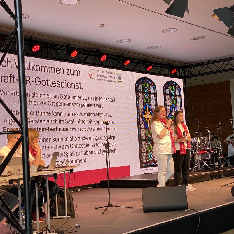 Zwei weiblich zu lesende Personen stehen auf einer Bühne und sprechen in Handmikrofone.