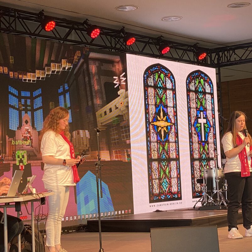 Zwei weiblich zu lesende Personen stehen auf einer Bühne. Eine der Personen spricht in eine Mikrofon.