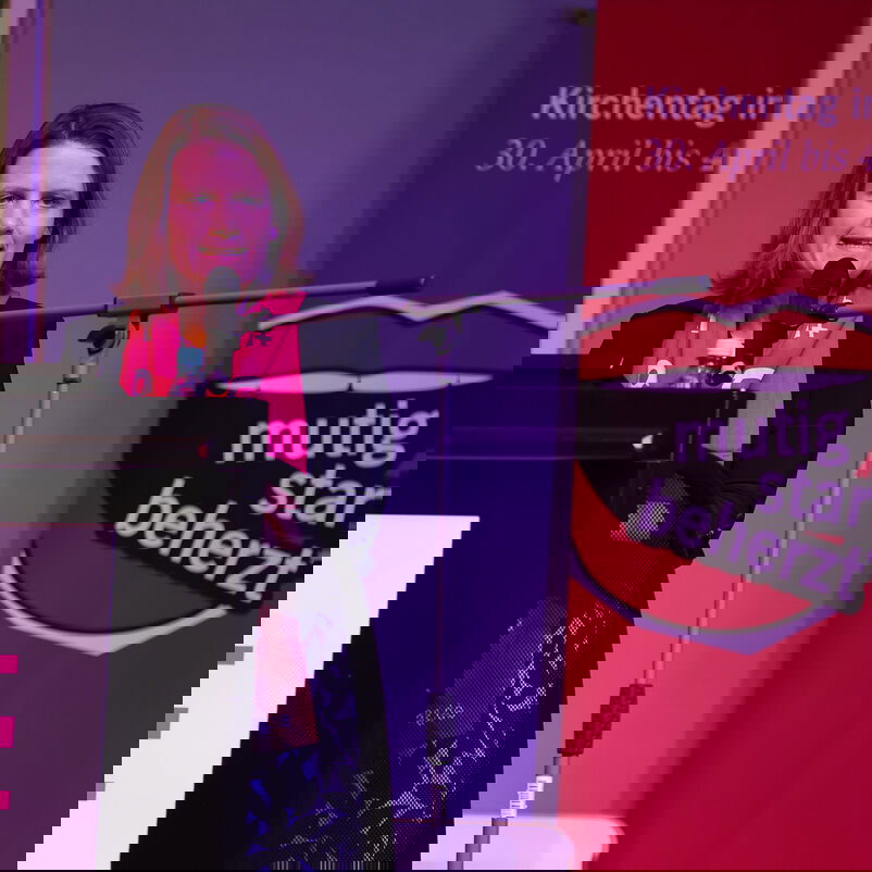 Eine als Frau lesbare Person steht an einem Redepult und spricht in ein Mikrofon.