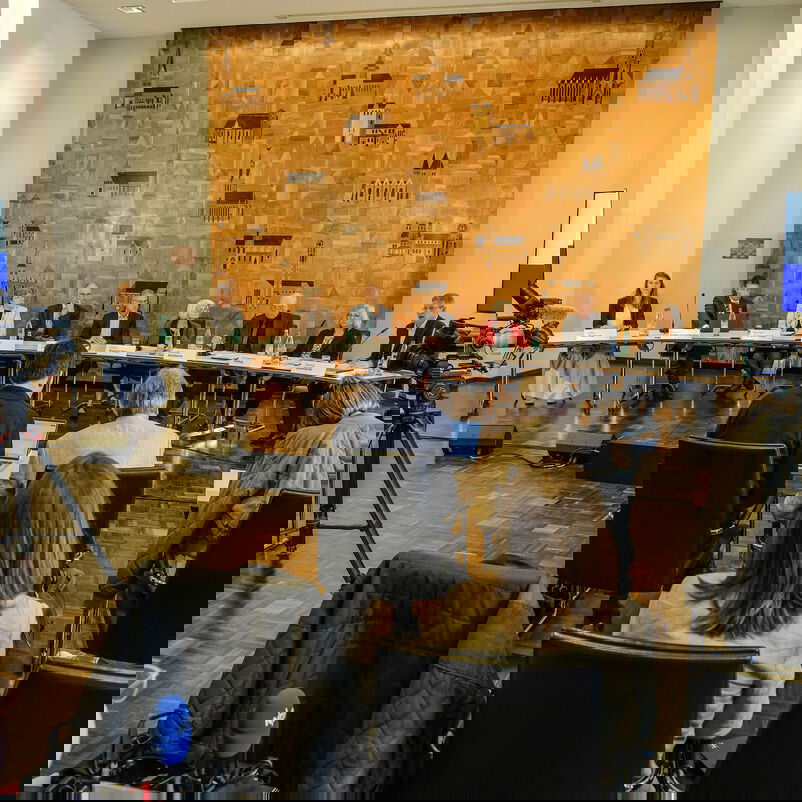 In einem Konferenzsaal sitzen eine Reihe von Menschen an Tischen, im Vordergrund stehen 