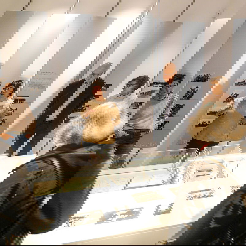 Weiblich und männlich zu lesende Personen stehen um einen Tisch in einem Museumsraum. Auf dem Tisch liegen Exponate.