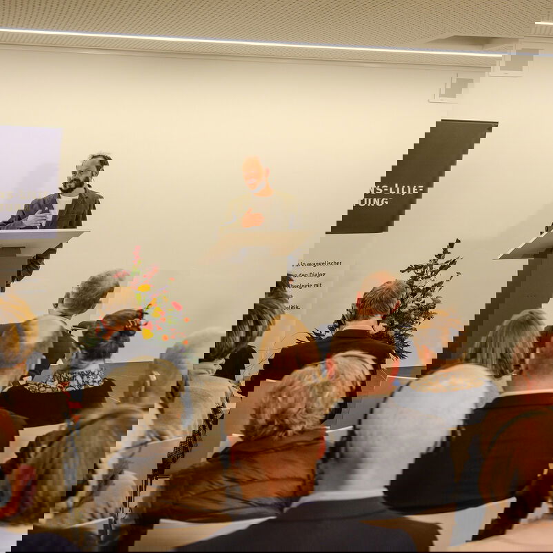 Eine männlich zu lesende Person steht an einem Redepult und spricht zu Menschen, die vor ihr sitzen.