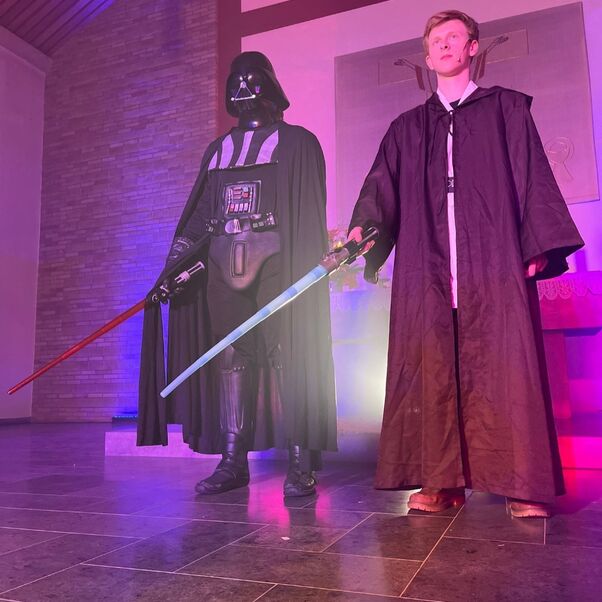 Man sieht jemanden in einem Darth-Vader-Kostüm. Daneben steht ein Jugendlicher mit Lichtschwert und dunklem Gewand als Luke Skywalker in einer Kirche. Die Kirche ist bunt beleuchtet.
