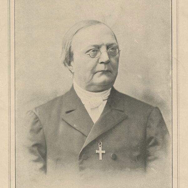 Das Bild zeigt eine Schwarzweiß-Aufnahme einer männlich lesbaren Person mit kurzen Haaren, einer Brille, einem schwarzen Sakko und einem weißen Hemd. Um den Hals hängt eine feine Kette mit einem Kreuz daran.