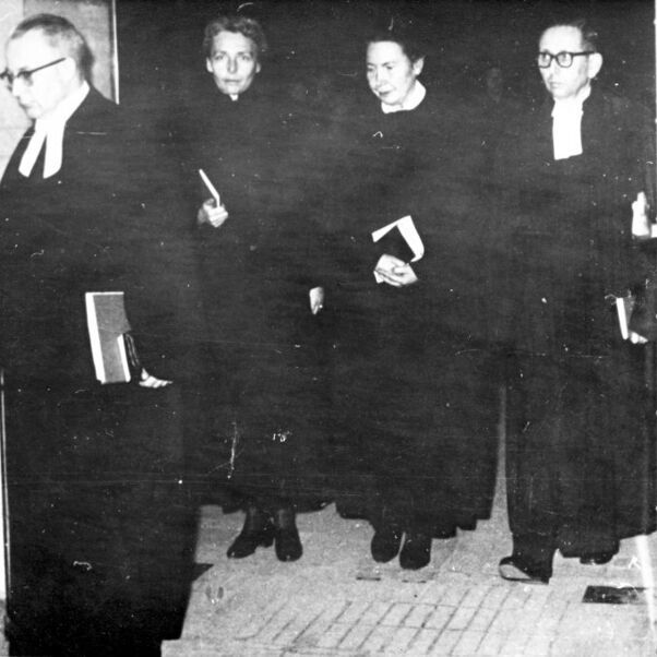 Das Schwarzweiß-Bild zeigt zwei männlich und zwei weiblich lesbare Personen in schwarzen Talaren, die gemeinsam aus einem Raum herauskommen.