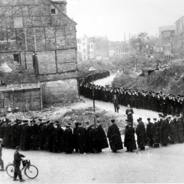 Das Schwarzweiß-Bild zeigt eine lange Reihe von schwarz gekleideten Menschen, die auf eine Kirche zustreben.