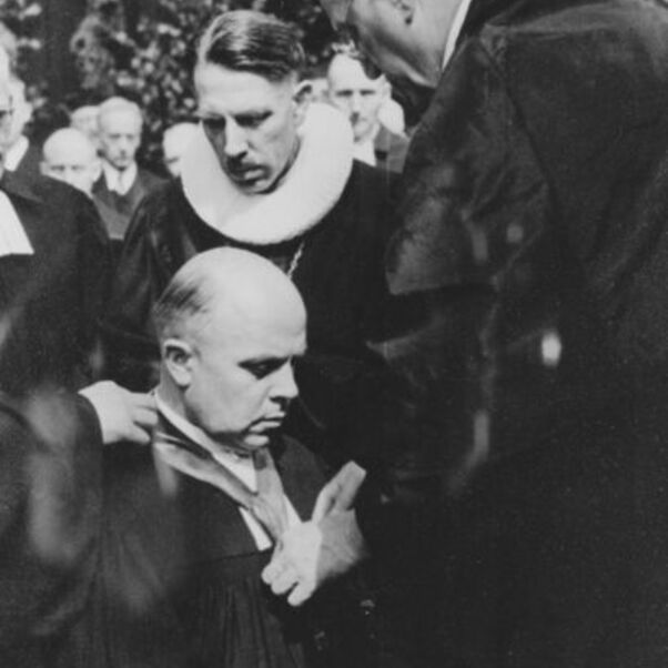 Das Bild zeigt eine knieende männlich lesbare Person, die eine schwere Kette zum Zeichen seiner Amtseinführung von einer anderen männlich lesbaren Person, beide in schwarzen Talaren,umgehängt bekommt.