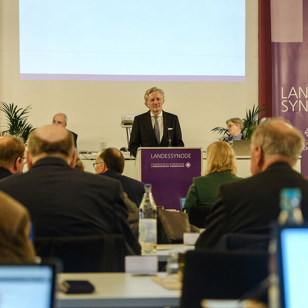 Eine männlich lesbare Person steht an einem Rednerpult vor einem Plenum