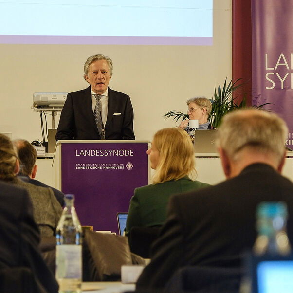 Eine männlich lesbare Person steht an einem Rednerpult vor einem Plenum