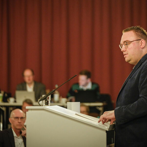 Eine männlich lesbare Person steht an einem Rednerpult vor einem Plenum