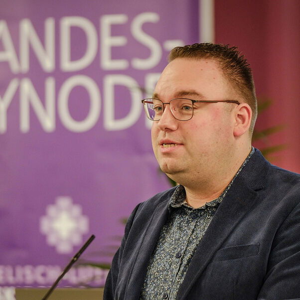 Eine männlich lesbare Person steht an einem Rednerpult vor einem Plenum