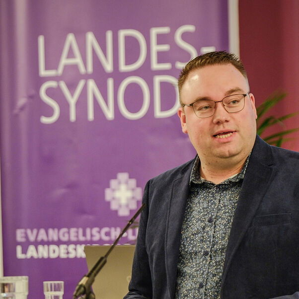 Eine männlich lesbare Person steht an einem Rednerpult vor einem Plenum