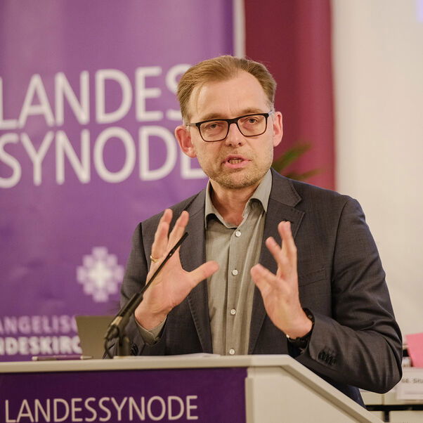 Eine männlich lesbare Person steht an einem Rednerpult vor einem Plenum