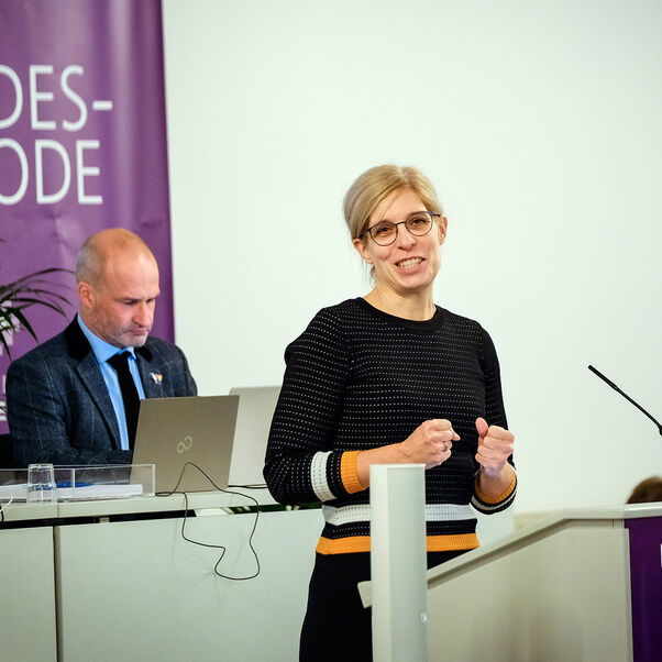 Eine weiblich gelesene Person mit zurückgebundenen blonden Haaren und Brille hinter einem Redepult