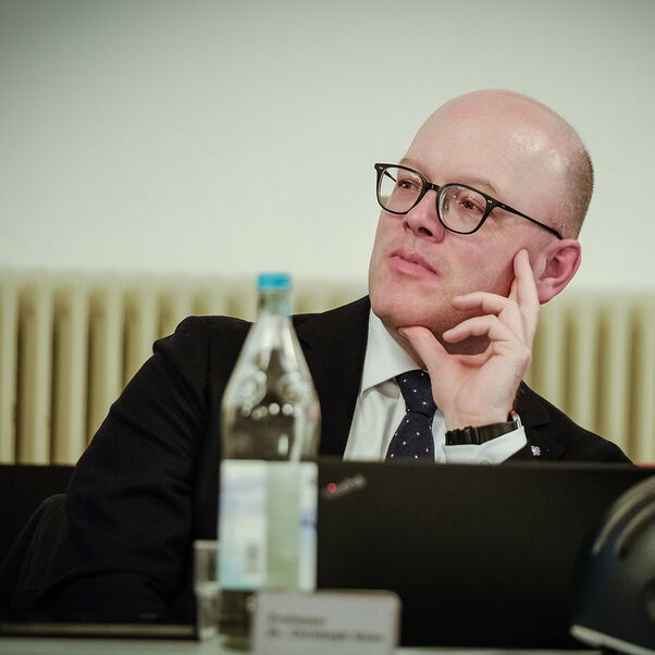 Eine männlich gelesene Person mit Brille sitzt am Tisch und hört zu. 