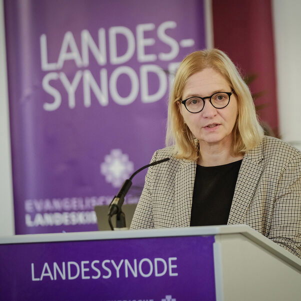 Eine weiblich gelesene Person mit schulterlangem blonden Haar und Brille hinter einem Redepult