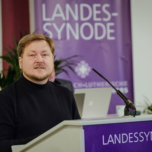 Eine männlich gelesene Person mit kurzen blonden Haaren und Vollbart hinter einem Redepult