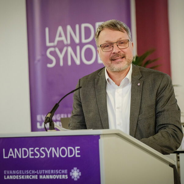 Eine männlich gelesene Person mit kurzen grauen Haaren und Brille hinter einem Redepult