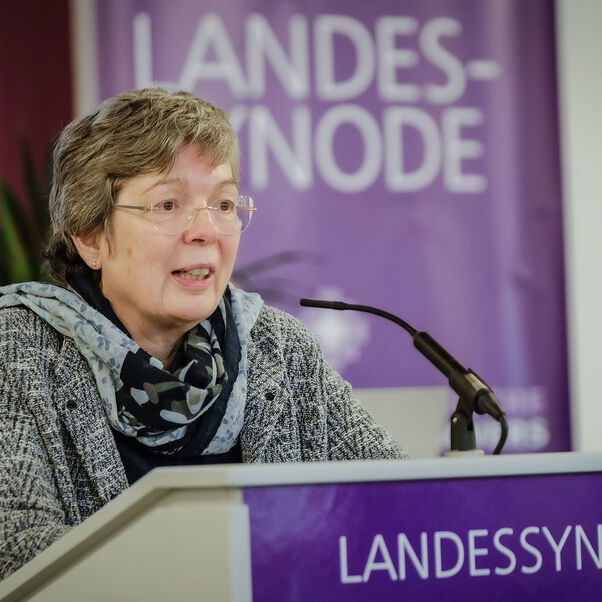 Eine weiblich gelesene Person mit kurzen grauen Haaren und Brille hinter einem Redepult.