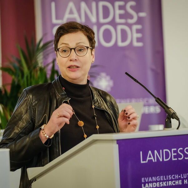 Eine weiblich gelesene Person mit kurzen dunklen Haaren und Brille hinter einem Redepult