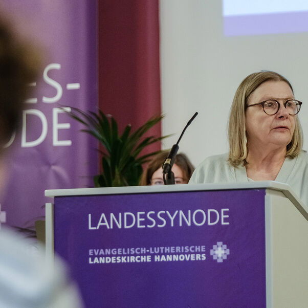 Eine weiblich gelesene Person mit schulterlangen blonden Haaren und Brille hinter einem Redepult