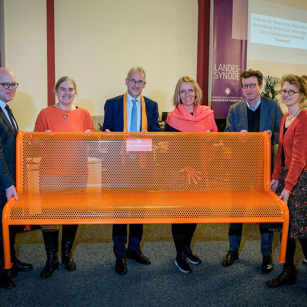 Sechs Personen, männlich und weiblich gelesene, halten gemeinsam eine große orange-farbene Bank aus Metall hoch. 