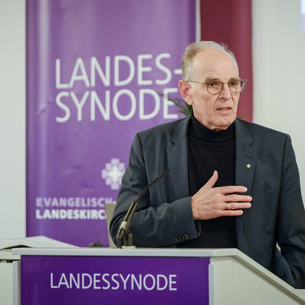 Eine männlich gelesene Person steht an einem Redepult im dunklen Jackett. 