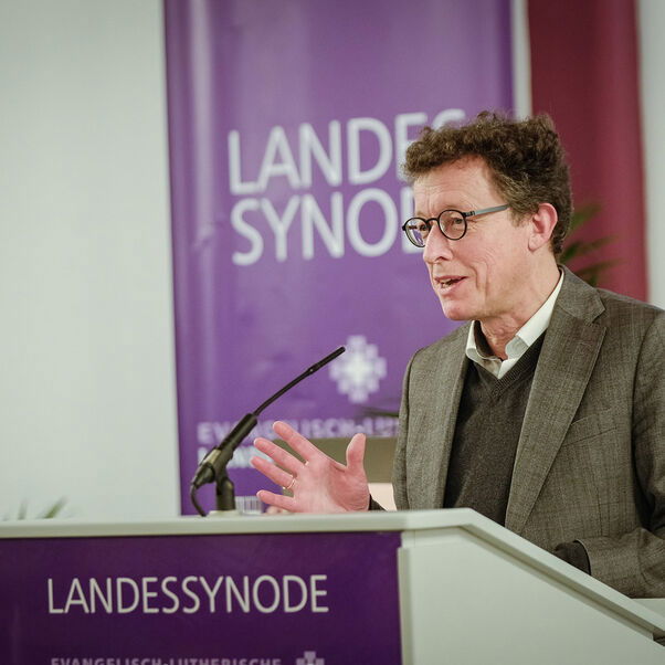 Eine männlich gelesene Person mit Brille und kurzen braunen Haaren im Jackett am Redepult. 