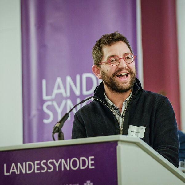 Eine männlich gelesene Person mit dunklen kurzen Haaren, Brille und Vollbart am Redepult. 
